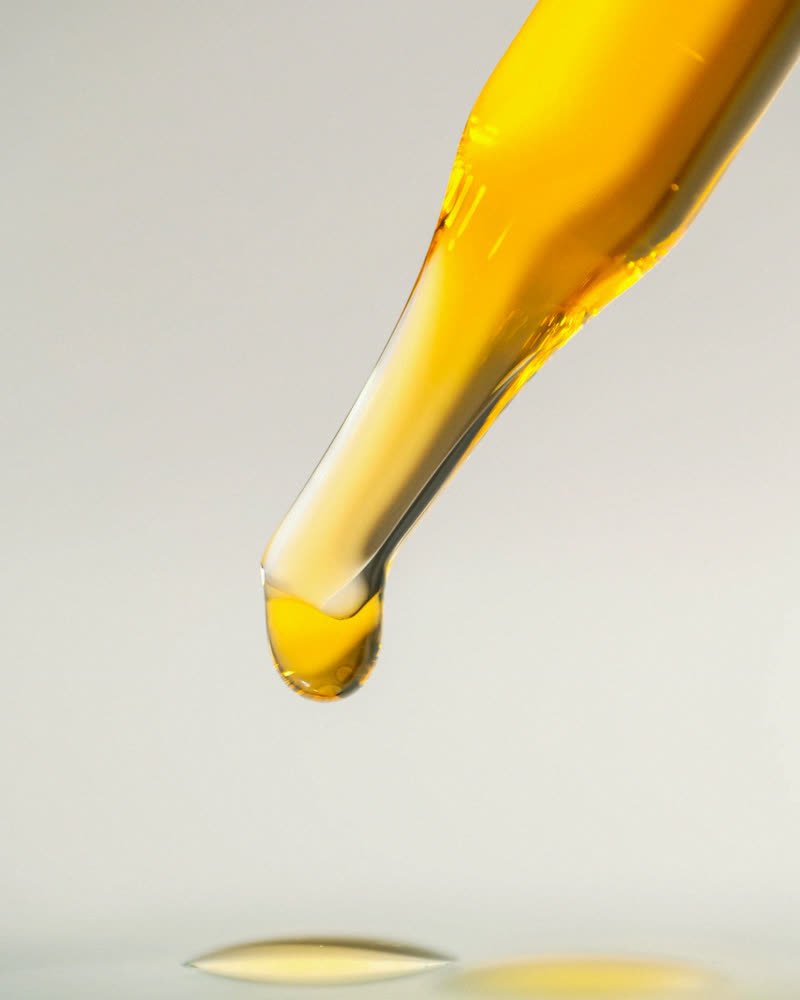 Uma gota de óleo âmbar se formando na ponta de uma pipeta de vidro, sobre fundo claro